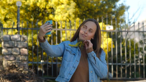 Pretty woman using smartphone and makes selfie against mimosa tree with flowers alt