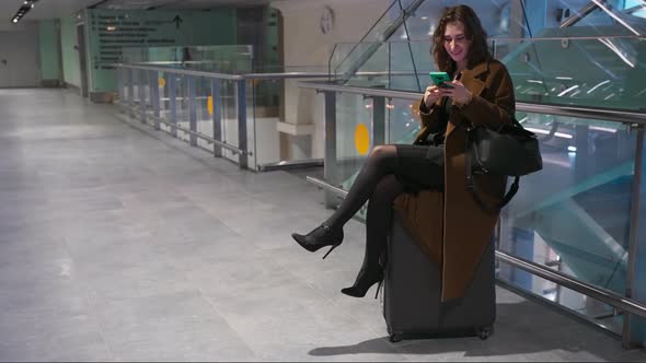 Bored Woman Girl is Sitting on a Suitcase and Uses a Smartphone Waiting in Airport Lounge Business