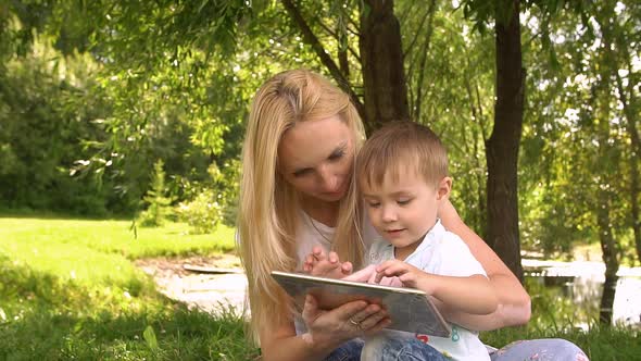 Happy Mother and Her Little Son Enjoying Playing on Tablet Computer in the Park alt