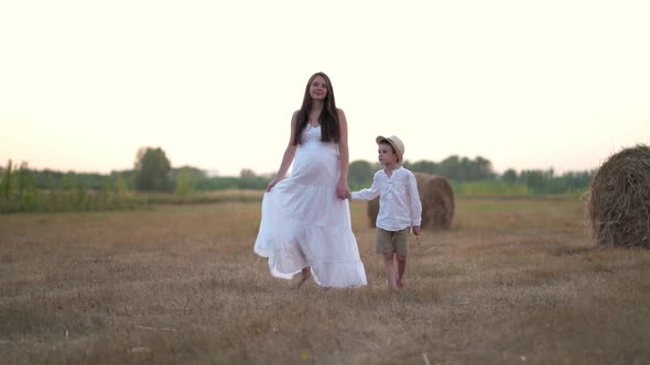 Beautiful Pregnant Woman with Son Walking in Wheat Field with Haystacks at Summer Day alt