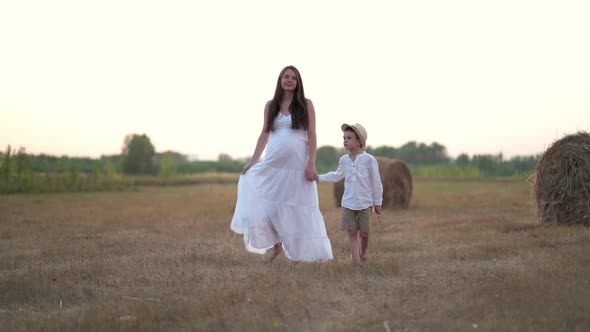 Beautiful Pregnant Woman with Son Walking in Wheat Field with Haystacks at Summer Day