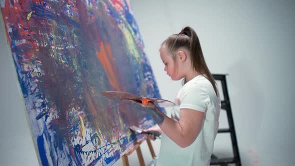 Girl with Special Needs Draws with a Brush on a Large Canvas in a White Room Kid Girl with Down alt