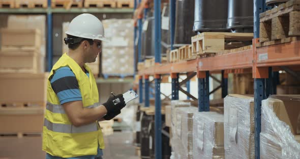 Logistics worker wearing a helmet working in a large warehouse checking inventory alt