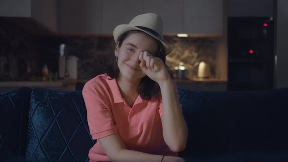 Girl in a Hat Watching a Movie at Night and Smiling Sitting on the Couch Background of the Kitchen alt