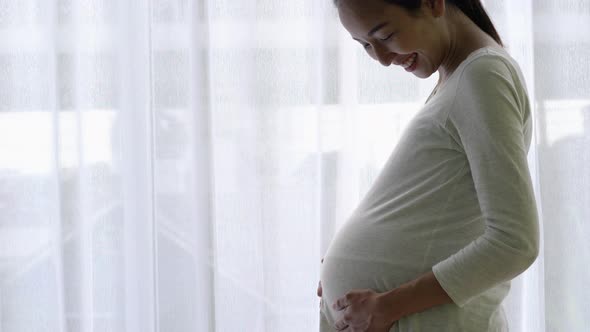 Pregnant woman looking through window and rubbing belly at home.