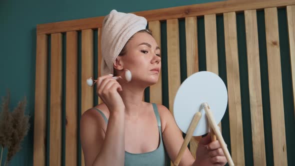Young Woman Using Jade Facial Roller for Face Massage Sitting on Bed in Bedroom Looking in the alt
