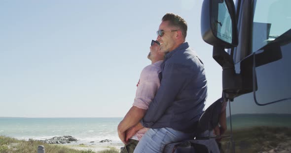 Happy caucasian gay male couple in sunglasses embracing, leaning on car on sunny day by the sea alt