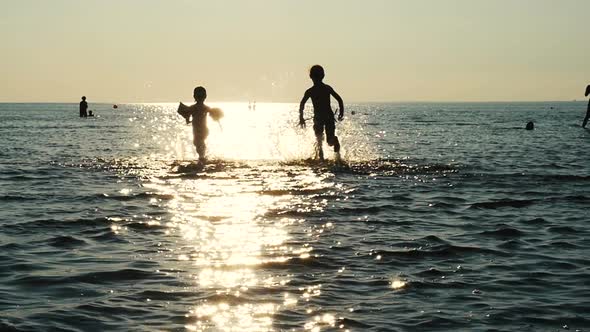 Children Run In The Sea Water In Sun Flare, Kids Silhouettes Running In The Sea. 