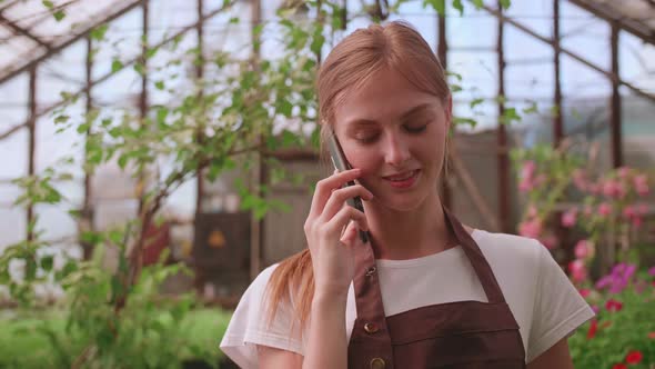 Young Girl Nursery Worker and Flower Greenhouse Speaks to a Client on the Phone