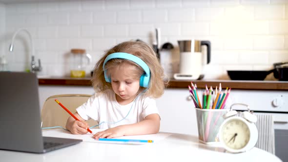 Little Girl in Headphones and in Front of a Laptop at Home in the Kitchen Draws Learning Online alt