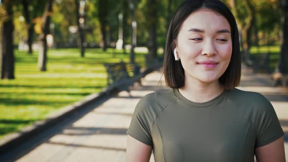 Adorable Asian Woman in Wireless Headphones Smiling to Camera in City Park Tracking Shot alt