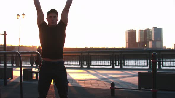 Young Sportive Man Squatting and Training His Hands with Dumbbells on Sunset alt
