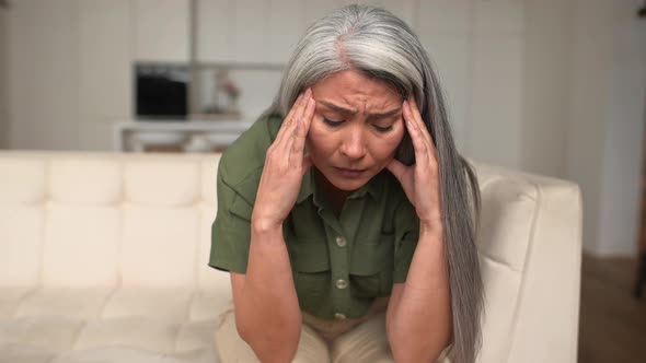 Grayhaired Woman Holding Forehead with Hand Sitting on the Sofa at Home alt