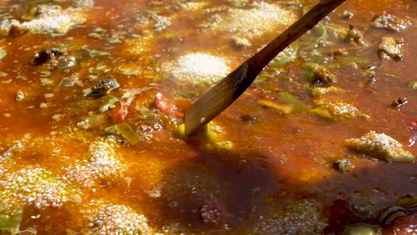 A close up shot of a wooden spoon mixing a soupy Spanish Paella while it is being cooked outside alt