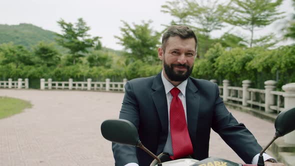 Portrait of a Senior Bearded Smiling Businessman Driving Up on a Motorcycle Along the Street alt