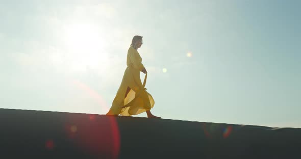 Stylish Woman in Yellow Dress Walking on Top of Sand Dune in Slow Motion alt