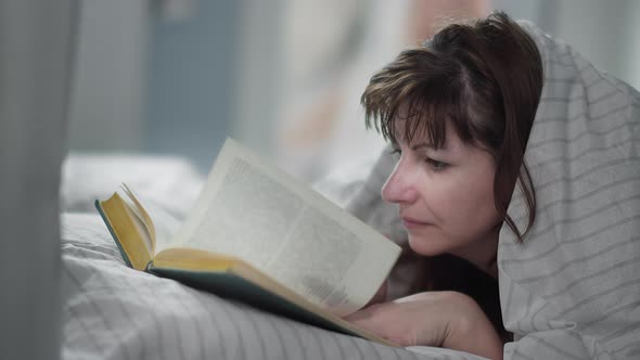 Woman Lies on the Bed and Reads a Book, Covered with a Blanket