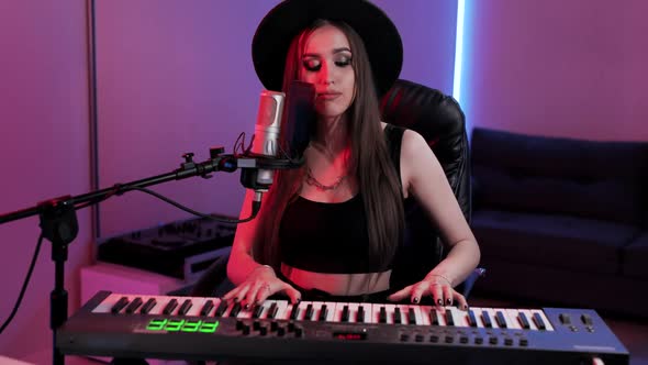 Beautiful Musician Woman in Hat Sings and Plays the Synthesizer at the Same Time in Recording Studio