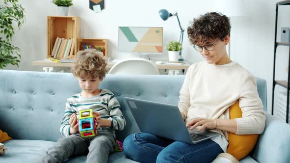 Businesswoman Working with Laptop Typing While Child Son Playing with Toy in Apartment alt