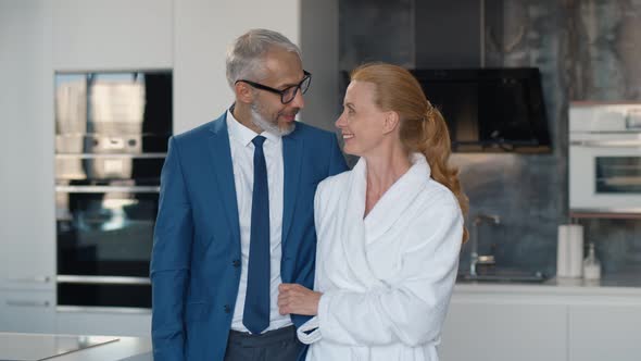 Handsome Mature Businessman in Suit and His Wife in Bathrobe Hugging in Home Kitchen alt