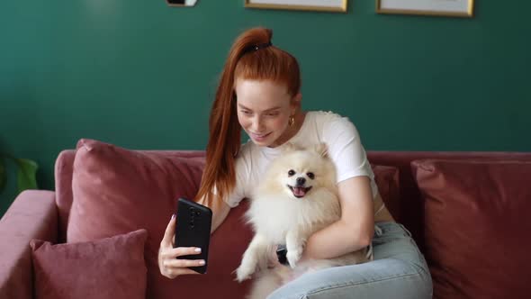 Cheerful Redhead Young Woman Hugging Pretty White Small Spitz Pet Dog and Taking Selfie Using Mobile alt