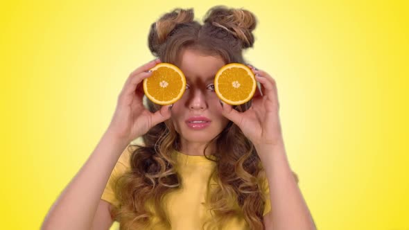 Attractive Girl in a Yellow Vest Closes Her Eyes with Oranges and Smiles Looking Into the Camera alt