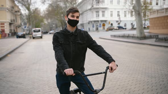 Portrait of Young Guy in Protective Face Mask Sitting on His Bike in the Street of Empty City Center alt