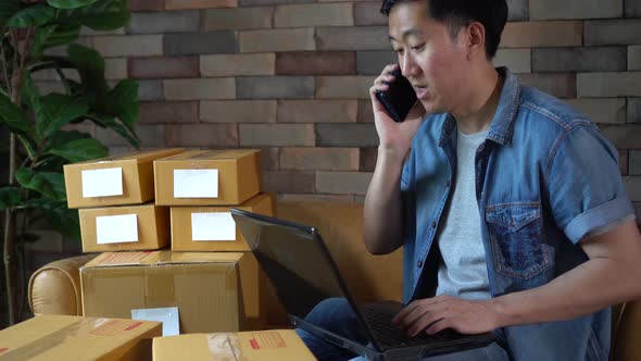 Asian Male Business Entrepreneur Using Laptop and Phone with Packs of Boxes at Home alt
