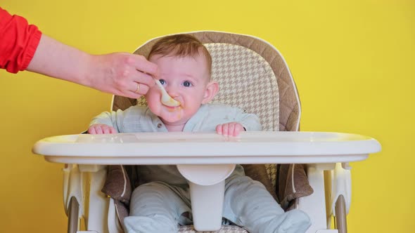 Mother feeding toddler baby from a spoon on a high chair for children, studio yellow background