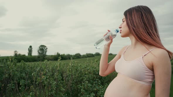 Pregnant young woman with dark hair in a pink top and light pants drinks water outdoors. 