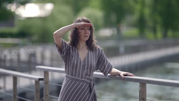 Portrait of a Brunette in a Striped Dress at the Railing of a Bridge Over the River on a Windy Sunny alt