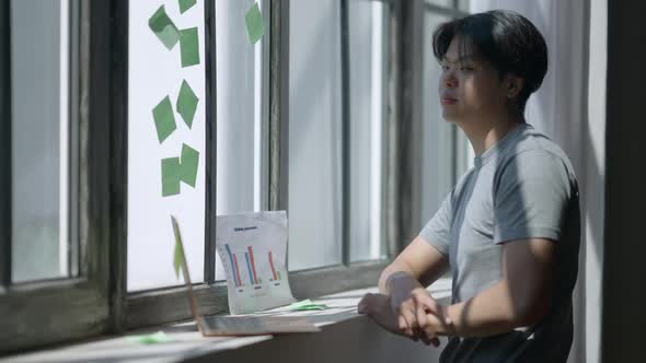 Young Thoughtful Asian Man Standing at Window in Home Office Planning Business Strategy alt