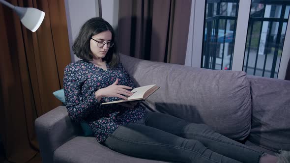 Girl in Glasses Sits on the Couch in the Evening with a Book Cars Drive Outside the Window alt