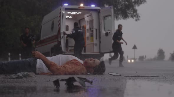 Team of Paramedics Helping Man Lying on Road Under Rain alt