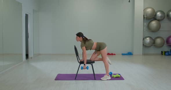 Sports Girl Performs Dumbbell Row to Belt with Emphasis on a Chair