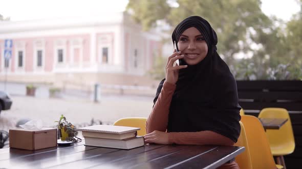 Young Pretty Muslim Woman in Hijab Talking on Phone and Smiling Sitting in Cafe Cheerful alt