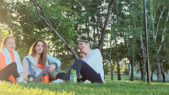 Three Girls Friends Are Sitting on the Grass After a Morning Jogging Exercise and Talking. alt