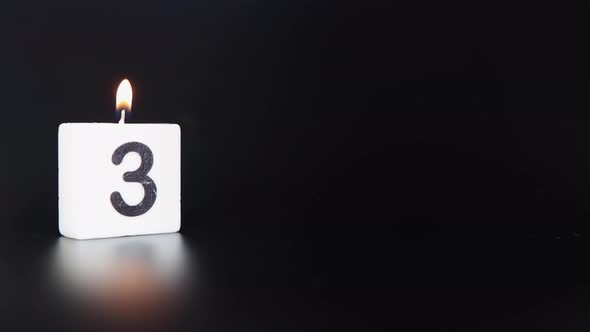 A square candle saying the number 3 being lit and blown out on a dark black background alt