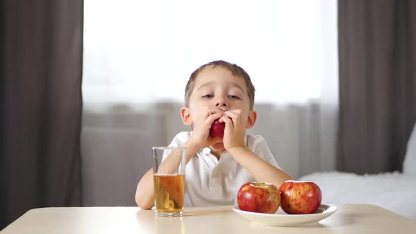 Happy Laughing Boy Sitting at the Table and Drinking Fruit Juice. The Child Bites Off a Slice of Red alt