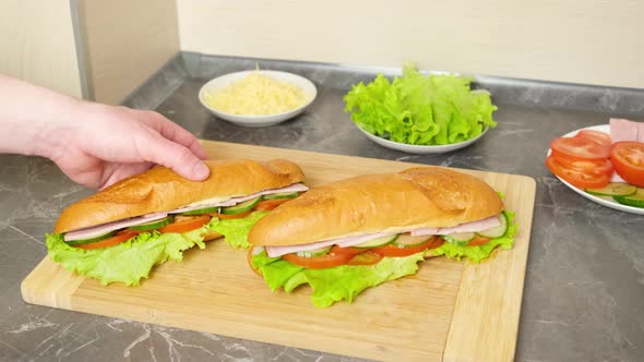Hands pick up freshly made sandwiches with vegetables, ham and cheese from a wooden cutting board.