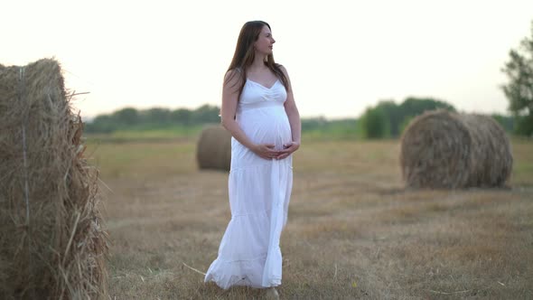 Beautiful Pregnant Woman in Wheat Field with Haystacks at Summer Day alt