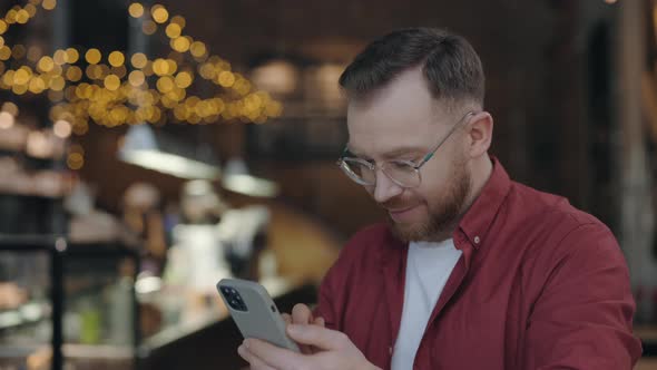 Cheerful Guy is Using Smartphone for Messaging While Working at the Cafe alt