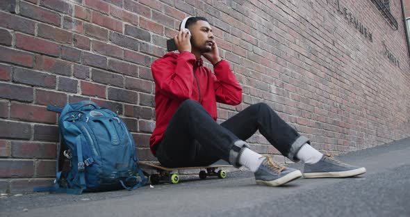 Mixed race man sitting on skateboard and listening to music in the street alt