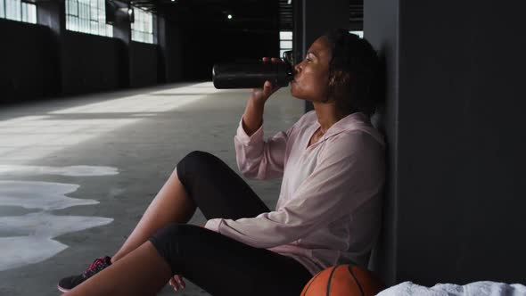 African american woman sitting in an empty building resting drinking water alt