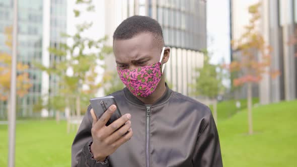 A Young Black Man in a Face Mask Reads Negative News on a Smartphone and Shakes His Head alt