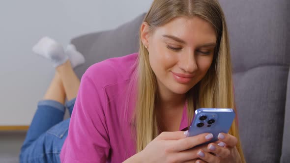Young blonde woman typing message on modern smartphone while lying on couch alt
