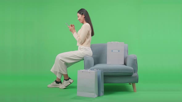 Woman With Shopping Bags Using Mobile Phone Shopping Online While Sitting On Sofa At Green Screen alt
