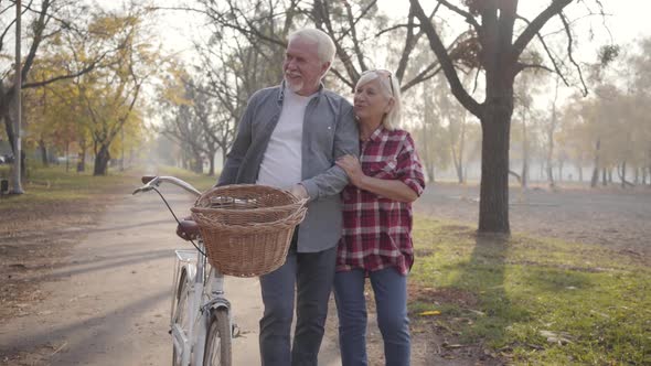 Happy Senior Caucasian Couple Walking with Bicycle Along the Alley in the Foggy Park alt