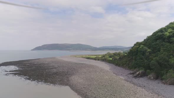Wide aerial tracking forward over a pebbly beach, beside a prehistoric looking forest cliff, reveali alt