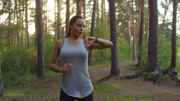 Cheerful Woman Running in Woods, Using Smartwatches and Posing for Camera alt